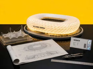 Maquetas arquitectónicas del Allianz Arena, el coloso en Munich y el Olympiastadion de Múnich sobre una mesa negra en una composición editorial deportiva.