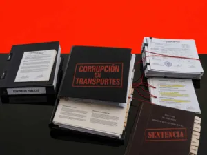 Expediente de investigación sobre corrupción en el Ministerio de Transportes, con documentos oficiales, archivos judiciales y contratos gubernamentales organizados sobre una mesa negra en composición editorial minimalista.